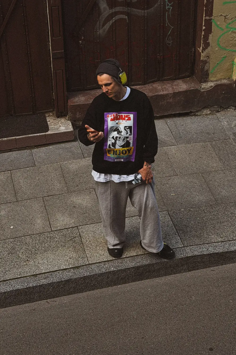 The figure standing on a stone sidewalk, looking down at a phone.